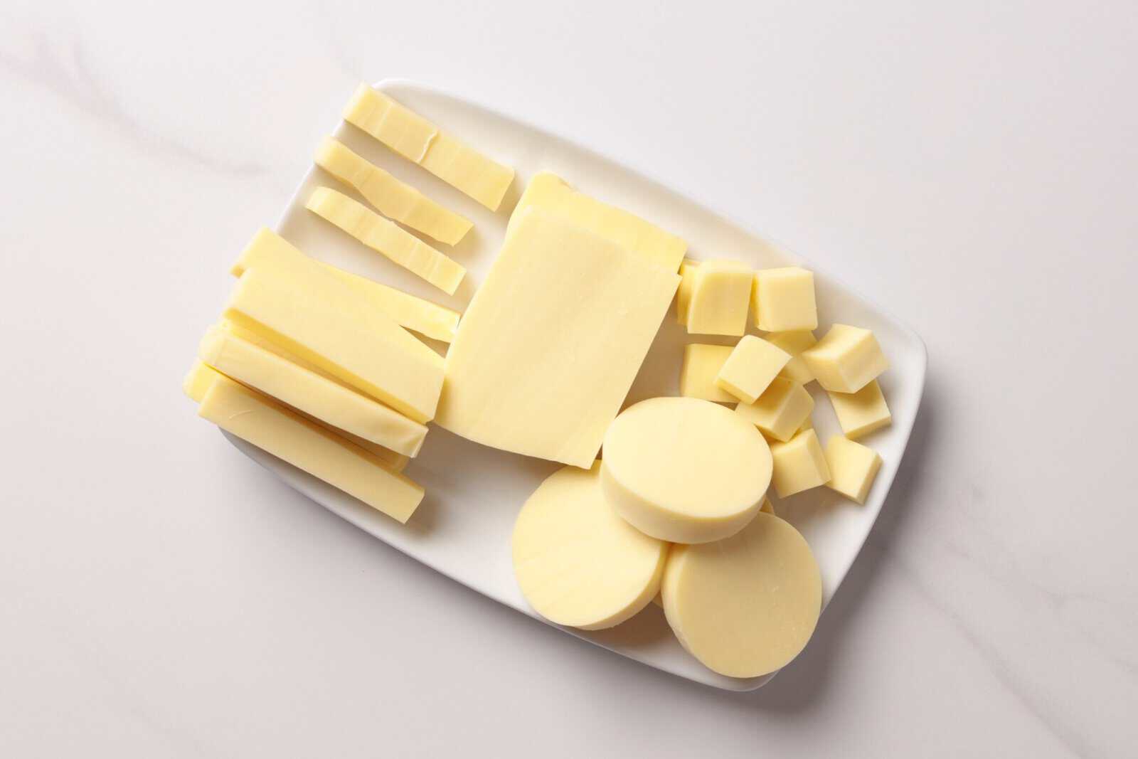 A white rectangular plate with sliced, cubed, and round pieces of mozzarella cheese arranged neatly on a light surface.