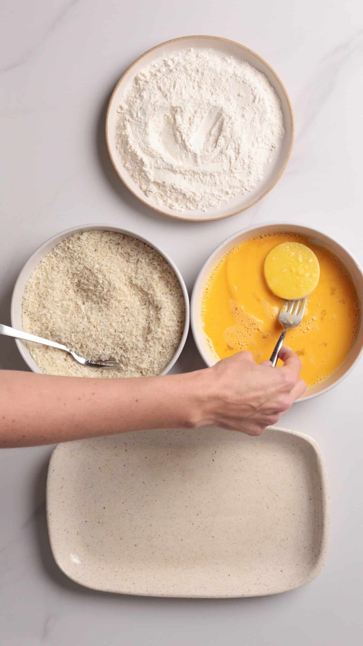 A hand dips a round mozarella slice into beaten eggs, next to bowls of flour and breadcrumbs, and an empty plate, all on a light countertop.