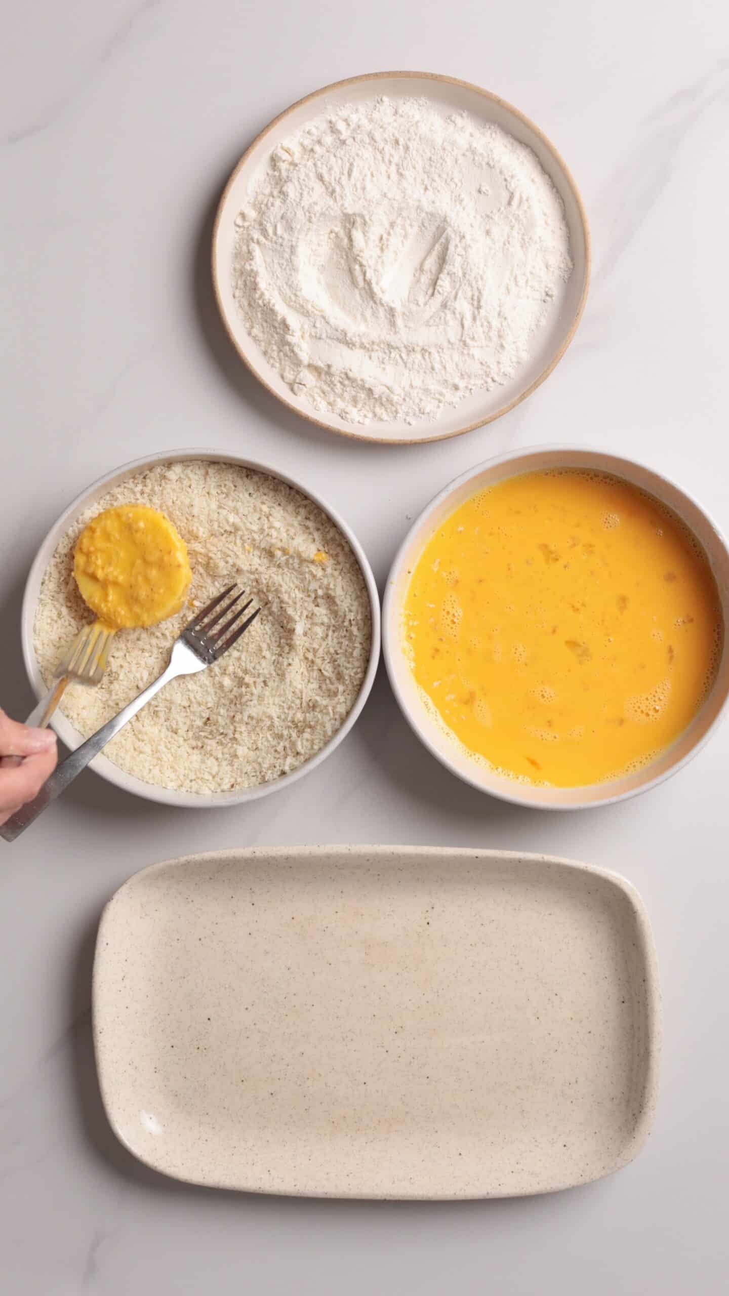 Four dishes: one with flour, one with beaten eggs, one with breadcrumbs, and one empty. A hand uses a fork to coat a mozzarella in breadcrumbs, showing a breading process on a white surface.
