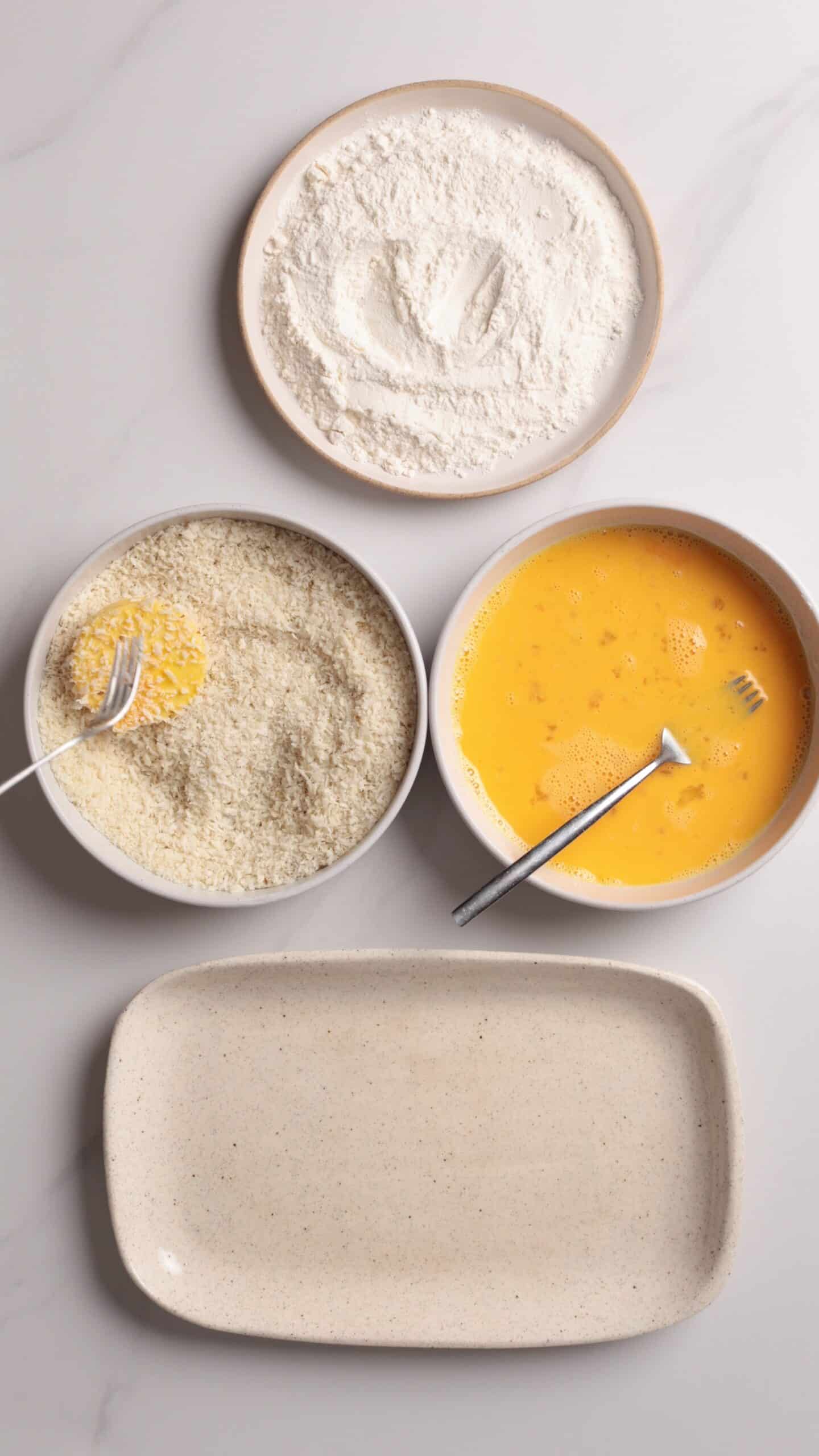 Three bowls with flour, beaten eggs, and breadcrumbs are arranged above an empty plate on a white surface. Forks are in the eggs and breadcrumbs bowls.