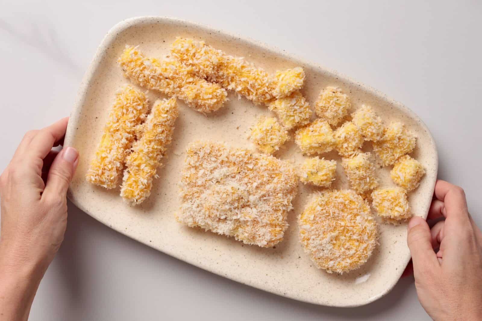 A person holds a speckled oval plate with various breaded, uncooked mozzarella cheese shapes including rectangles, cylinders, and a circle, on a white surface.