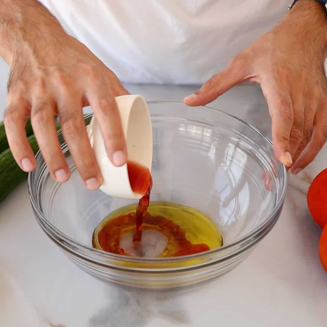 A person pours a red wine vinegar from a white cup into a glass bowl containing oil on a marble counter, with tomatoes and a cucumber nearby.