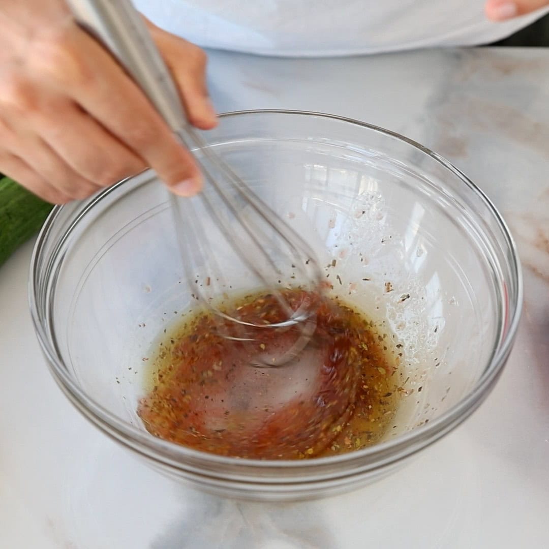 A person whisking a Greek salad dressing which contains oil, red wine vinegar, and seasonings in a glass bowl on a marble countertop.