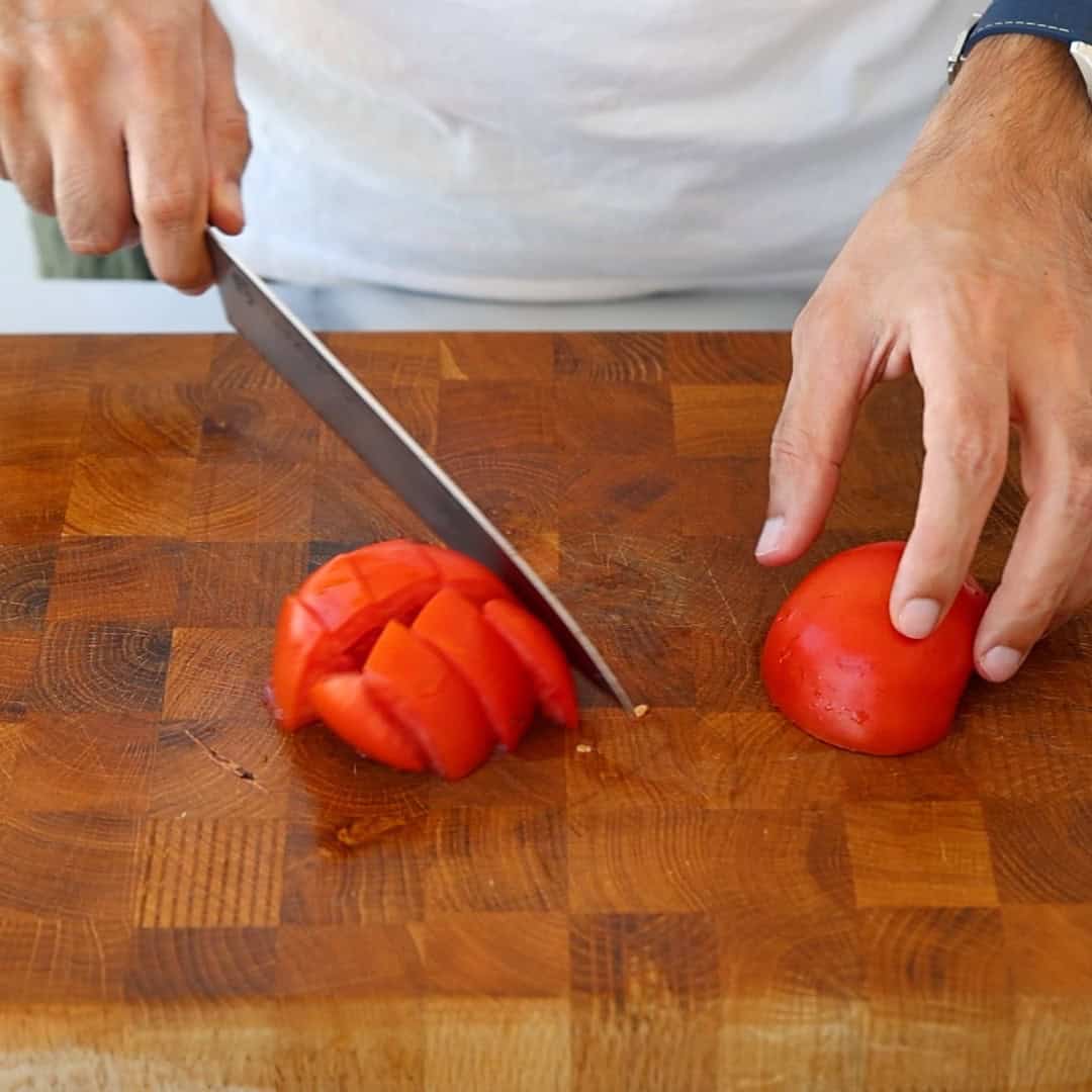 A person slices a red tomato on a wooden cutting board with a large knife, wearing a white shirt and a watch on their left wrist.