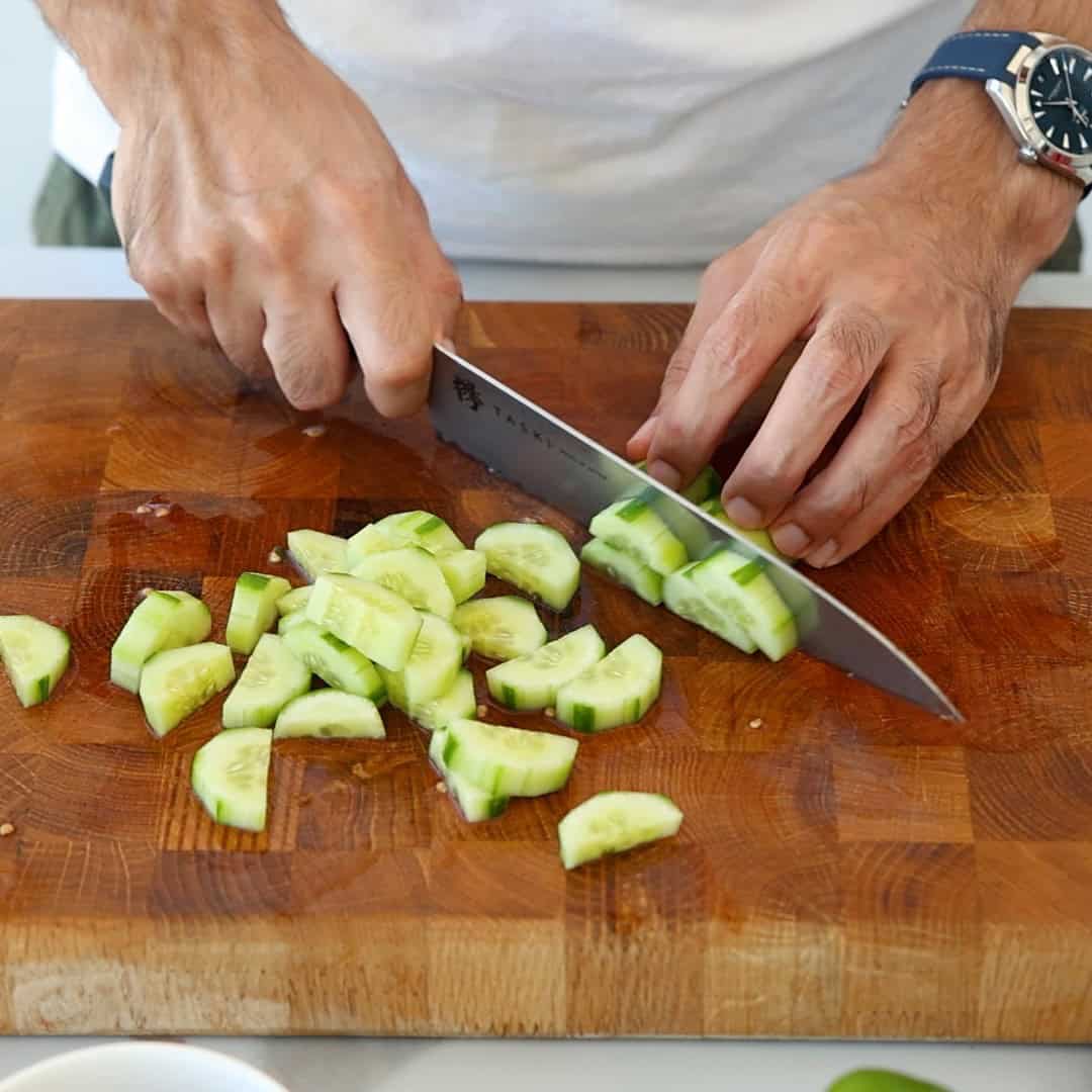 A person slices a cucumber on a wooden cutting board with a large knife. The cucumber pieces are scattered on the board, and a watch is visible on the person’s wrist.