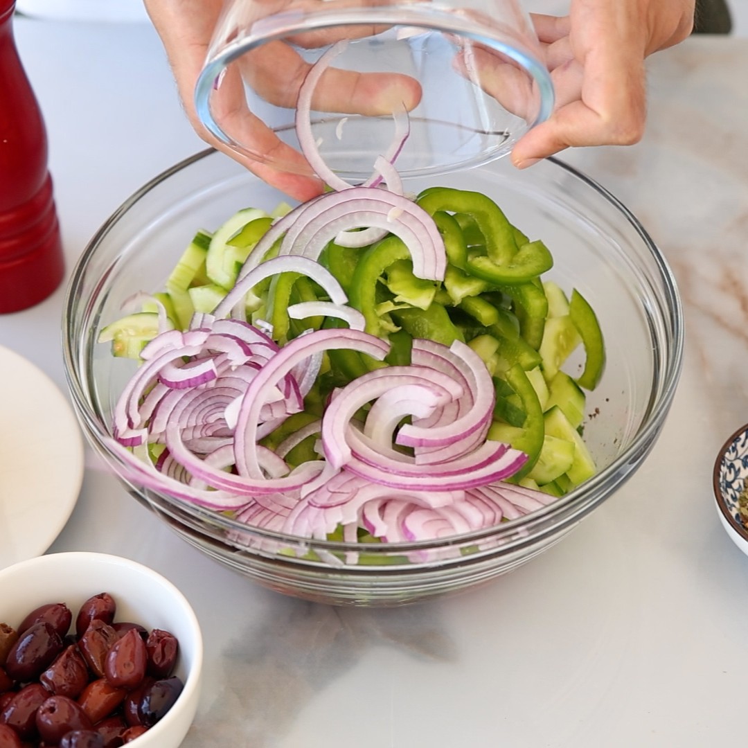 A person adds sliced red onions from a glass bowl into a larger bowl filled with chopped green bell peppers. A small bowl of black olives and a red pepper grinder are nearby on the counter.