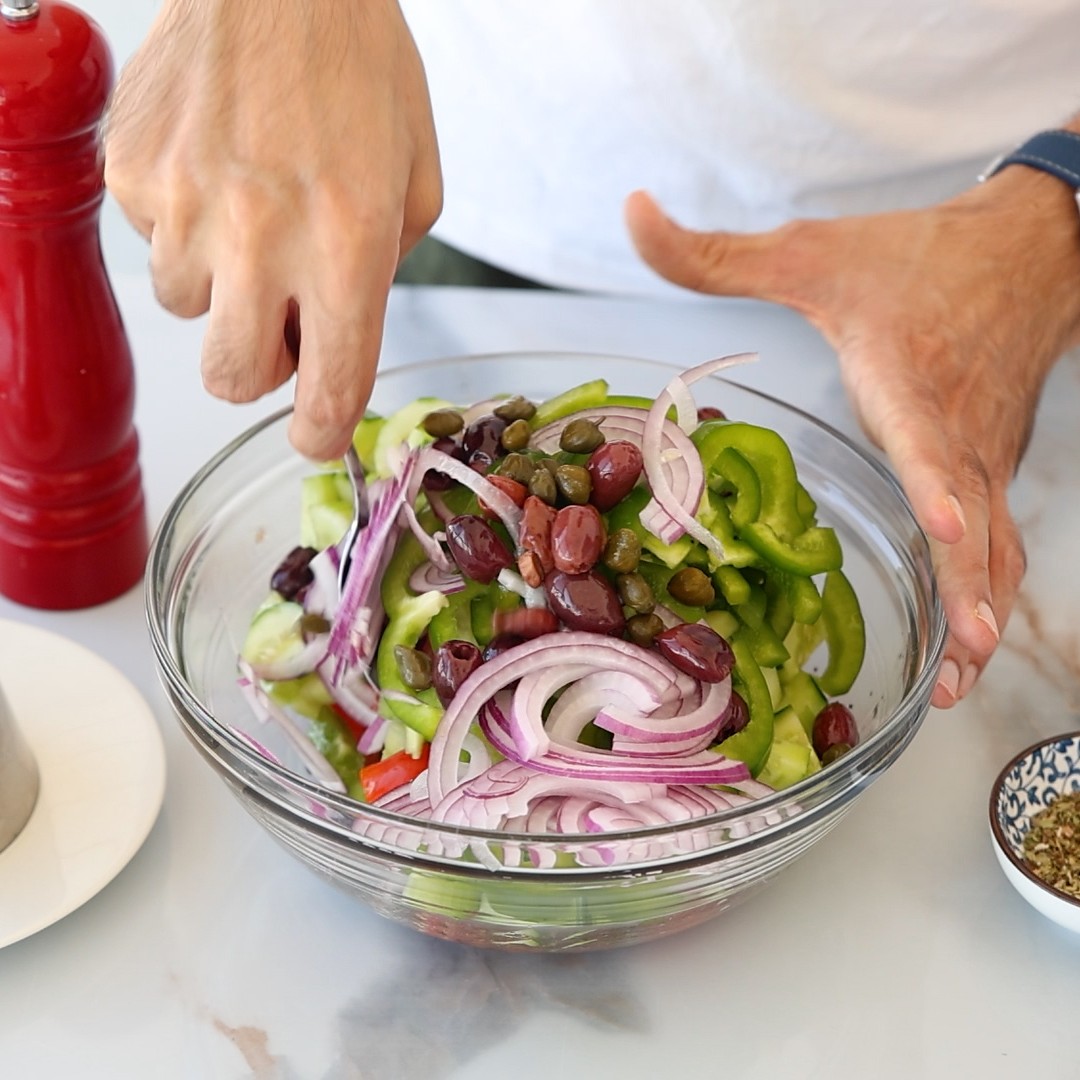 A person mixing a bowl of authentic horiatiki salad with sliced red onions, green peppers, cucumbers, olives, and capers on a countertop next to a red pepper grinder.