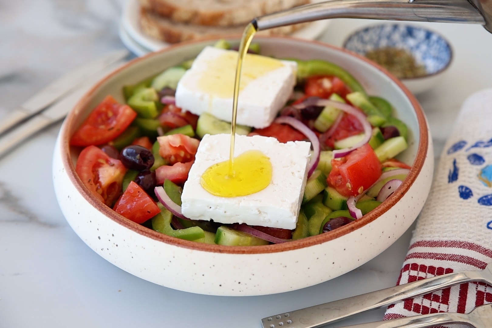 A Greek salad with tomatoes, cucumber, olives, red onions, and two feta slices is drizzled with olive oil. Sliced bread and a napkin are beside the bowl on a white surface.