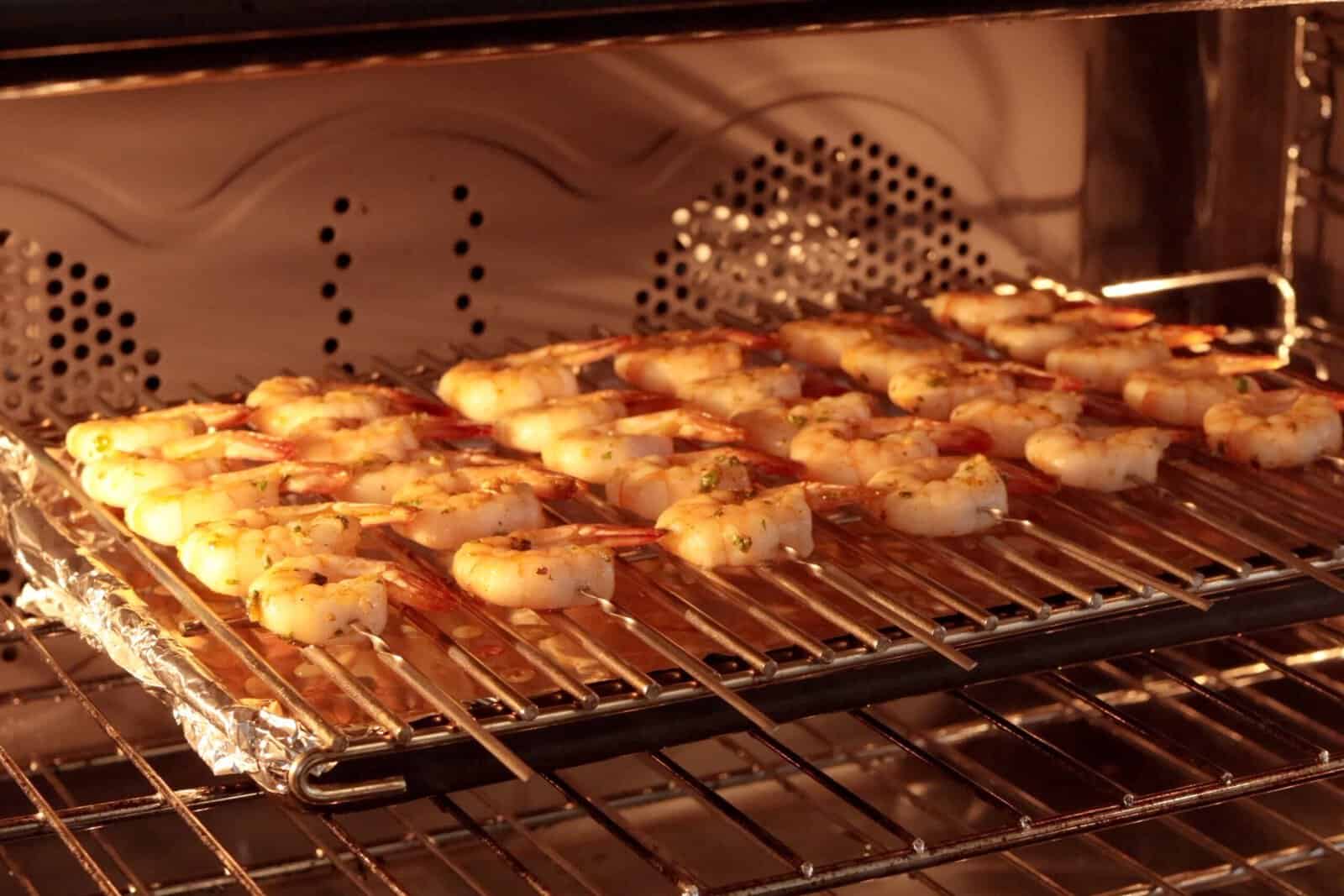 Shrimp seasoned with herbs and spices are arranged in rows on a wire rack inside an oven, with foil lining the oven tray beneath to catch drips. The shrimp are roasting or broiling.