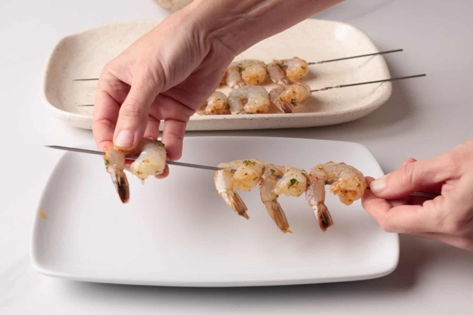 Hands threading marinated shrimp onto metal skewers over a white plate, with more shrimp skewers visible on a nearby rectangular dish in the background.