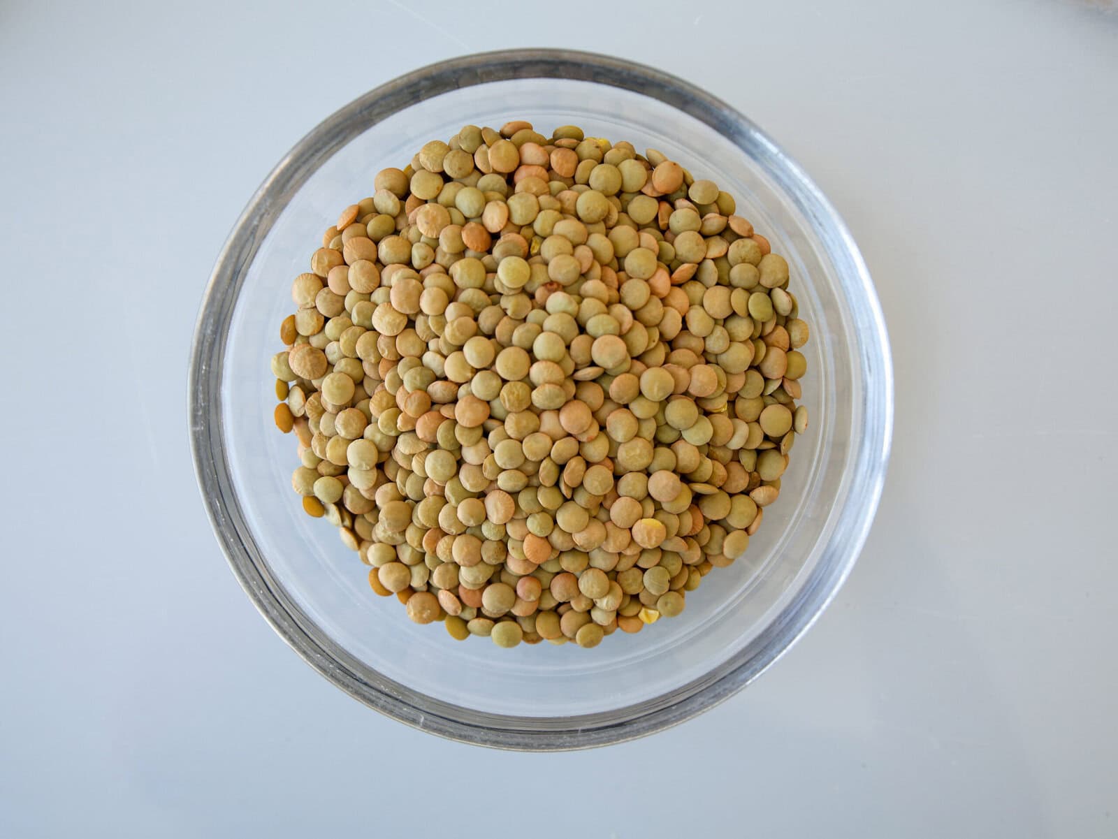 A clear glass bowl filled with dry green and brown lentils, viewed from above on a light gray surface.