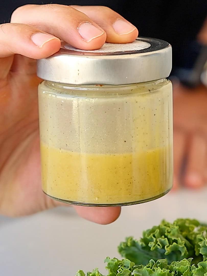 A hand holds a glass jar with a silver lid, containing a two-layer sauce—yellow on the bottom and creamy beige on top. Kale leaves are visible at the bottom right corner.