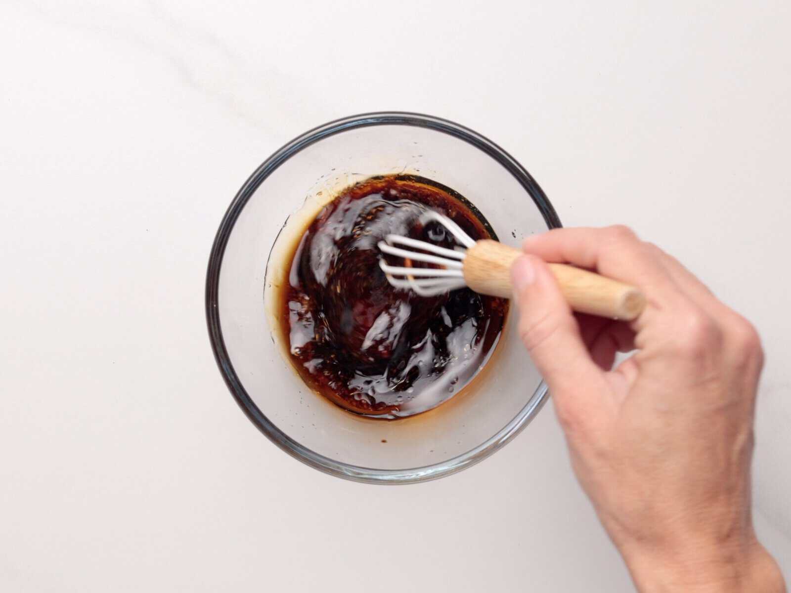 A hand whisking a marinade for tuna poke bowl in a clear glass bowl on a white surface.