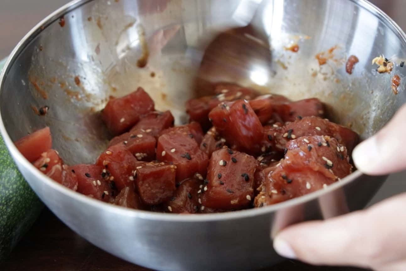 A hand holds a metal bowl filled with marinated tuna cubes, topped with sesame seeds. The bowl is on a wooden surface, and part of a cucumber is visible on the side.