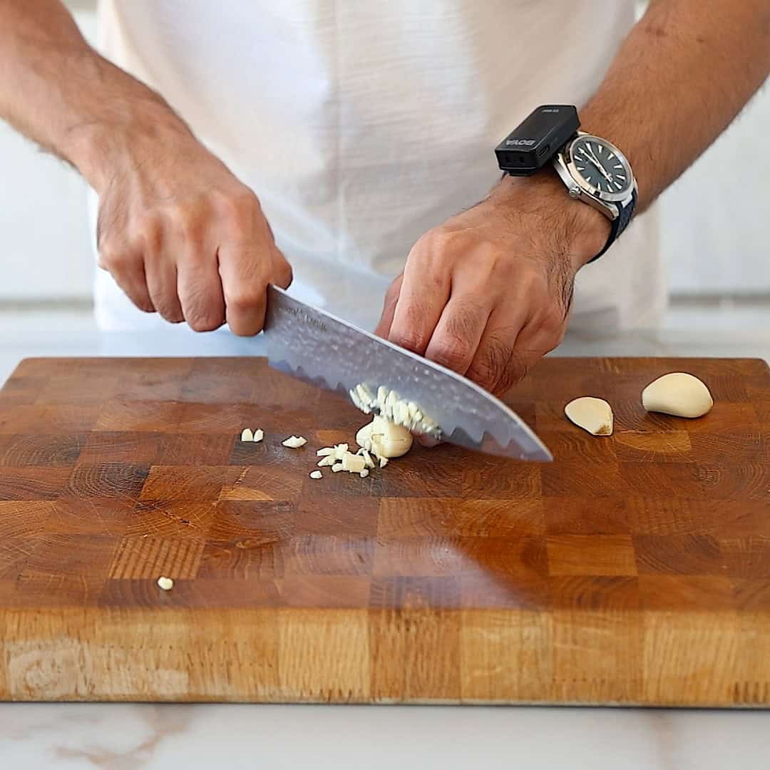 A person chops garlic cloves on a wooden cutting board with a large knife. They wear a white shirt, a wristwatch, and a small black device clipped to their chest.