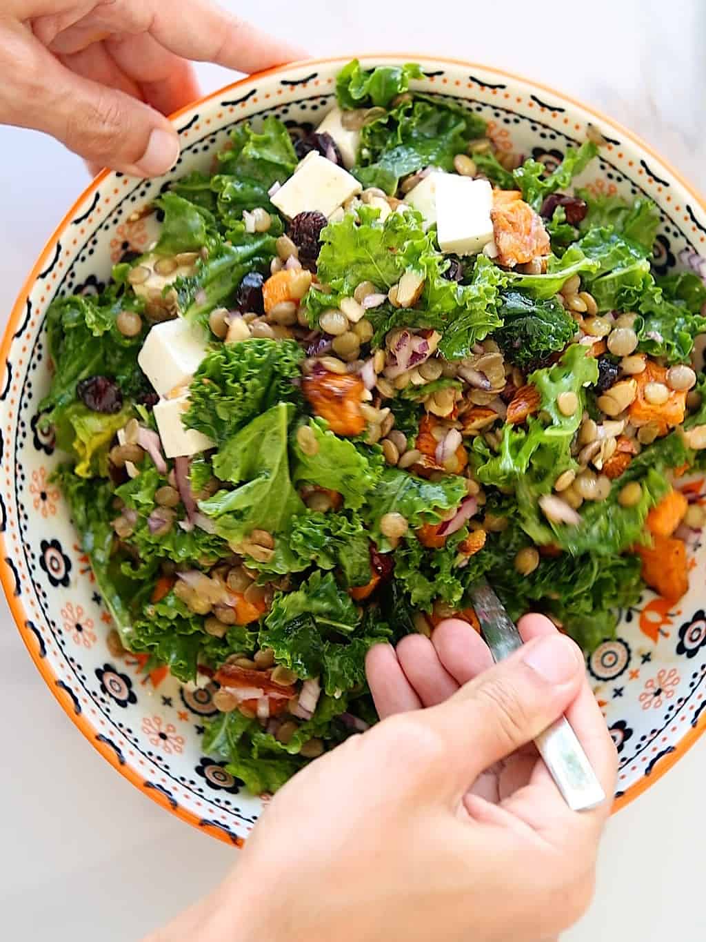 A person holding a colorful bowl of kale salad with grains, dried fruits, nuts, and cheese cubes, mixing it with a fork.