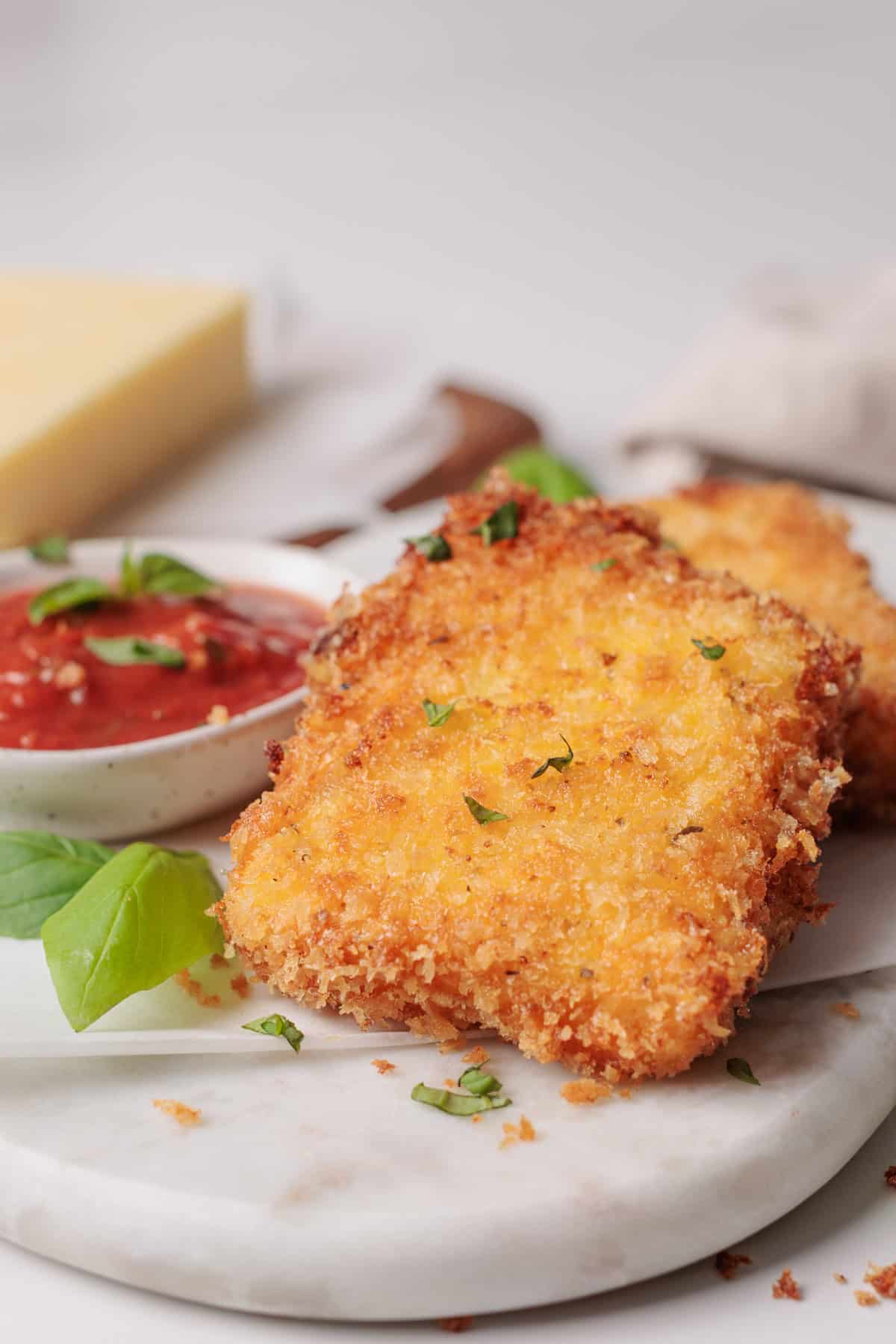Two golden, crispy, breaded fried mozzarella squares sit on a white plate with marinara sauce for dipping. Fresh basil leaves and a wedge of cheese are in the background.