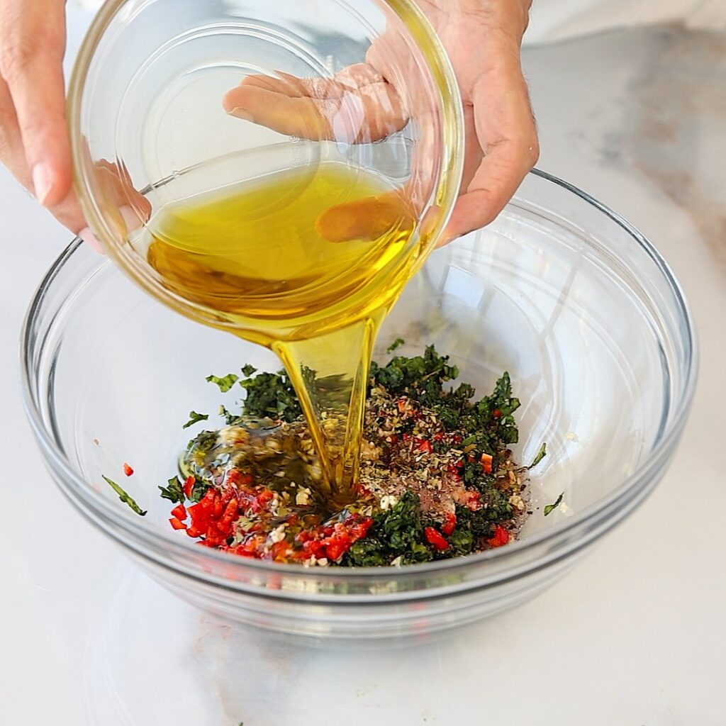 A person pours olive oil from a glass bowl into a larger bowl containing chopped herbs, red peppers, and spices on a white surface.