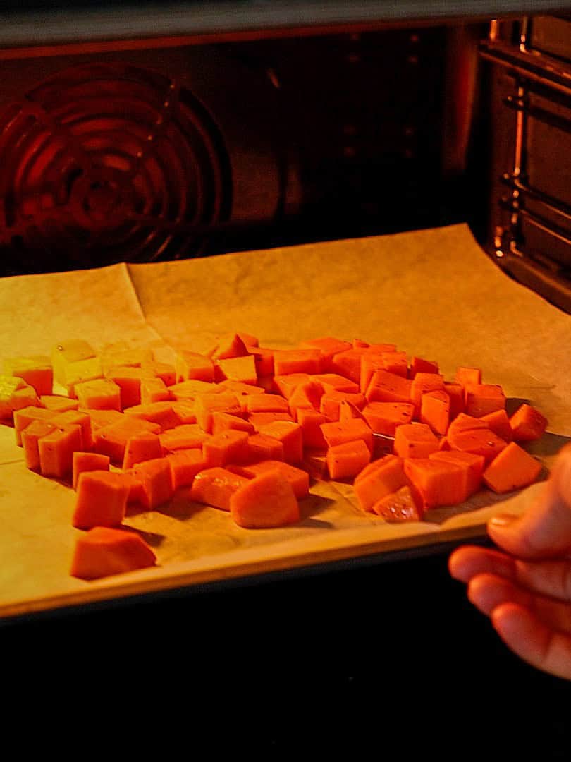 Diced sweet potatoes on a parchment-lined baking sheet are being placed into an oven for roasting.