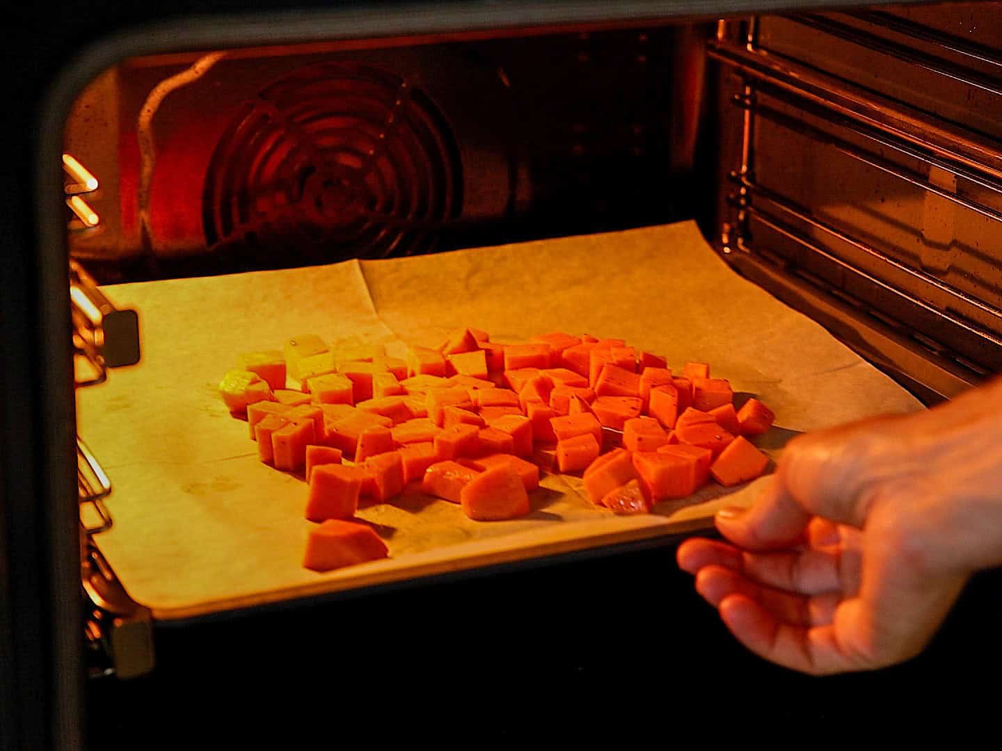 A hand places a baking tray with diced sweet potatoes on parchment paper into an oven for roasting.