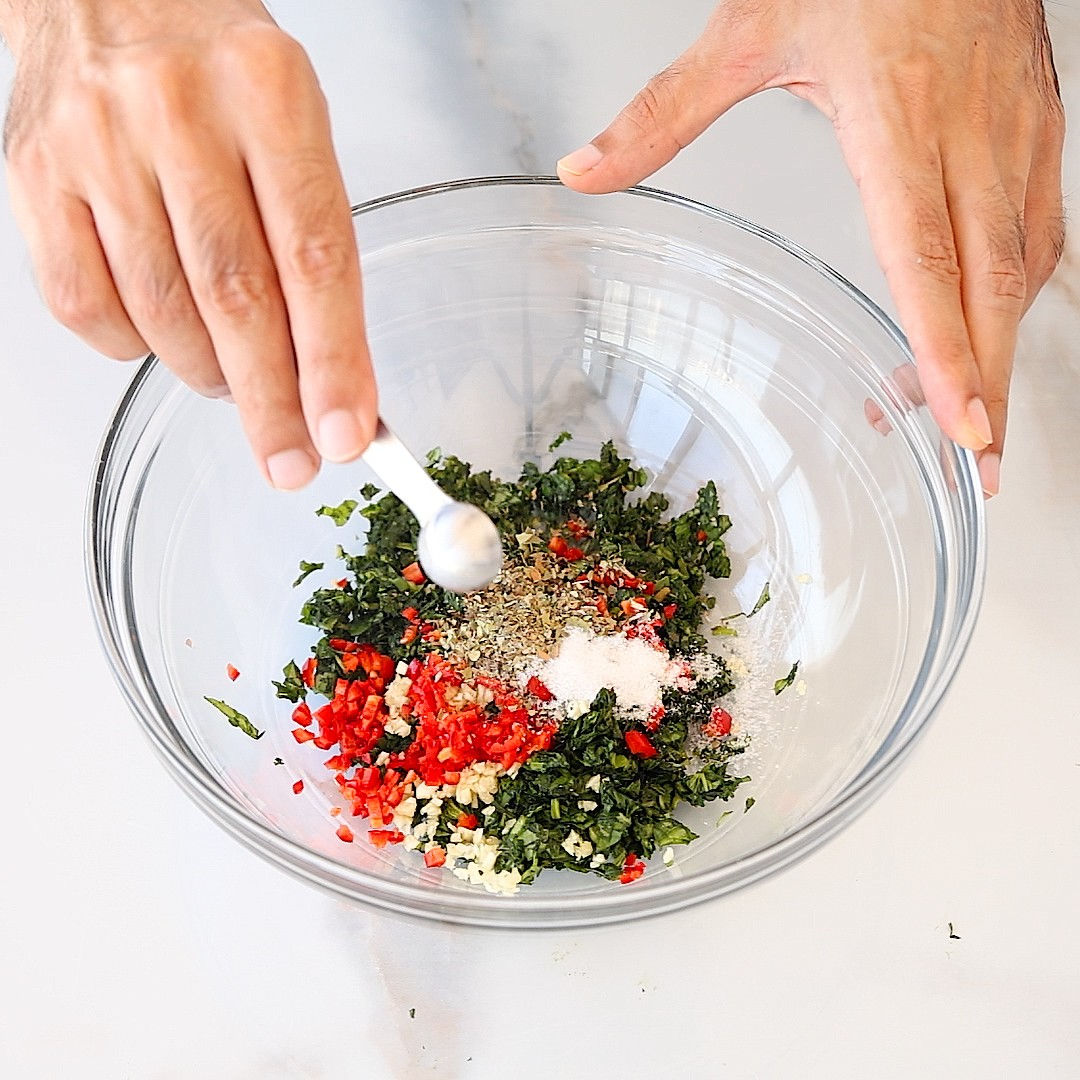 A person sprinkles seasoning from a spoon into a glass bowl containing chopped herbs, red chili, and garlic on a white countertop.