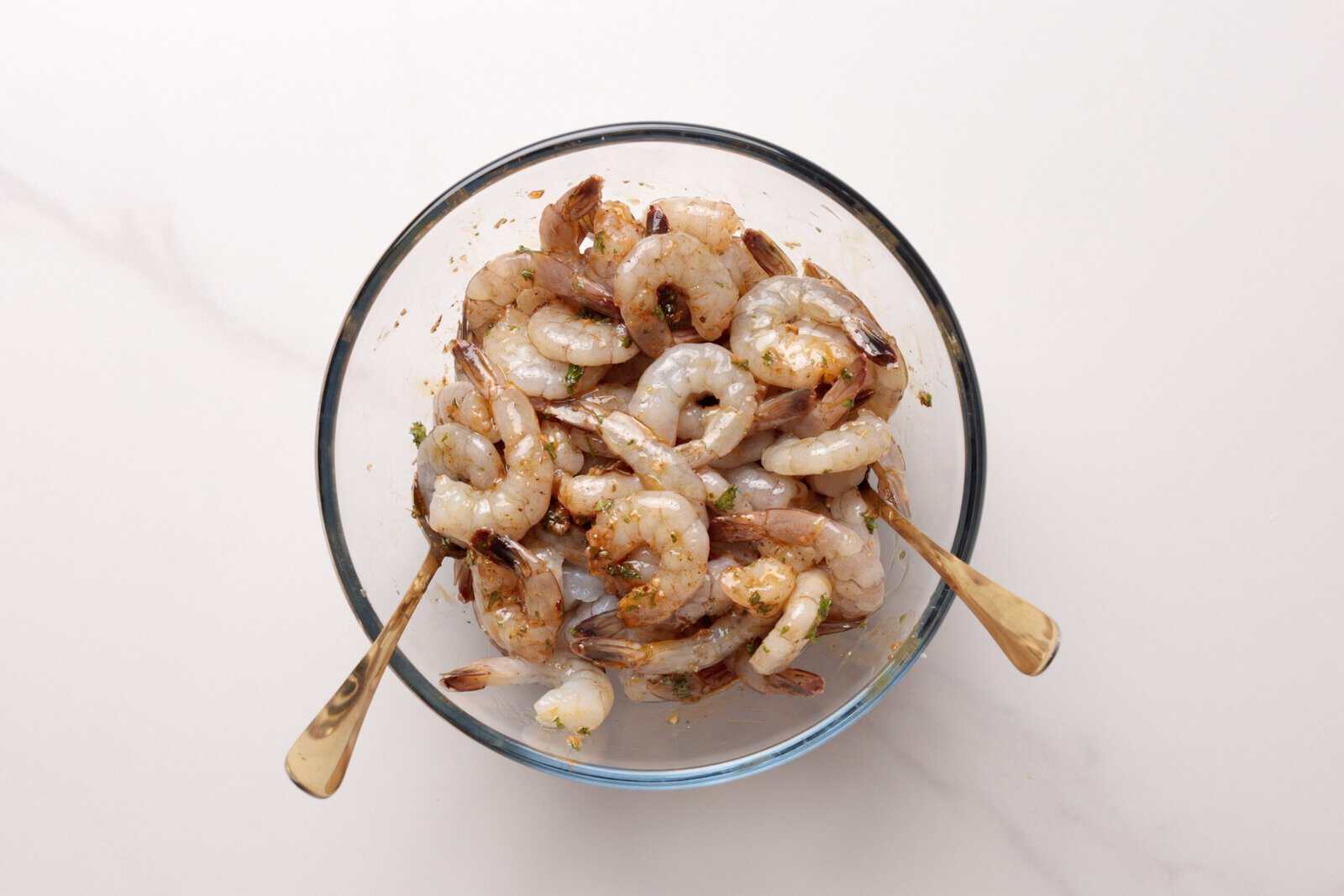 A glass bowl filled with raw, deveined shrimp mixed with seasoning. Two gold-colored forks are placed inside the bowl, and the background is a plain white surface.