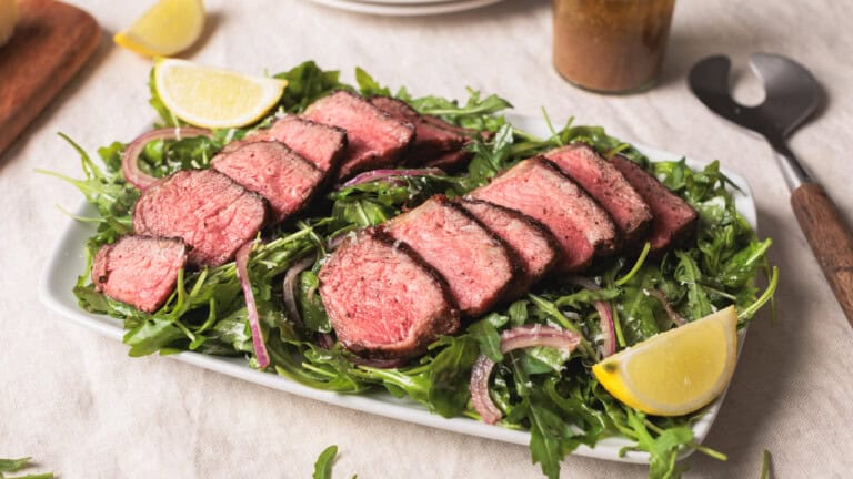 A rectangular plate with Steak salad with arugula. A rare steak on a bed of arugula and red onion, garnished with lemon wedges. A serving utensil, dressing bottle, and lemon slices are nearby on a light tablecloth.