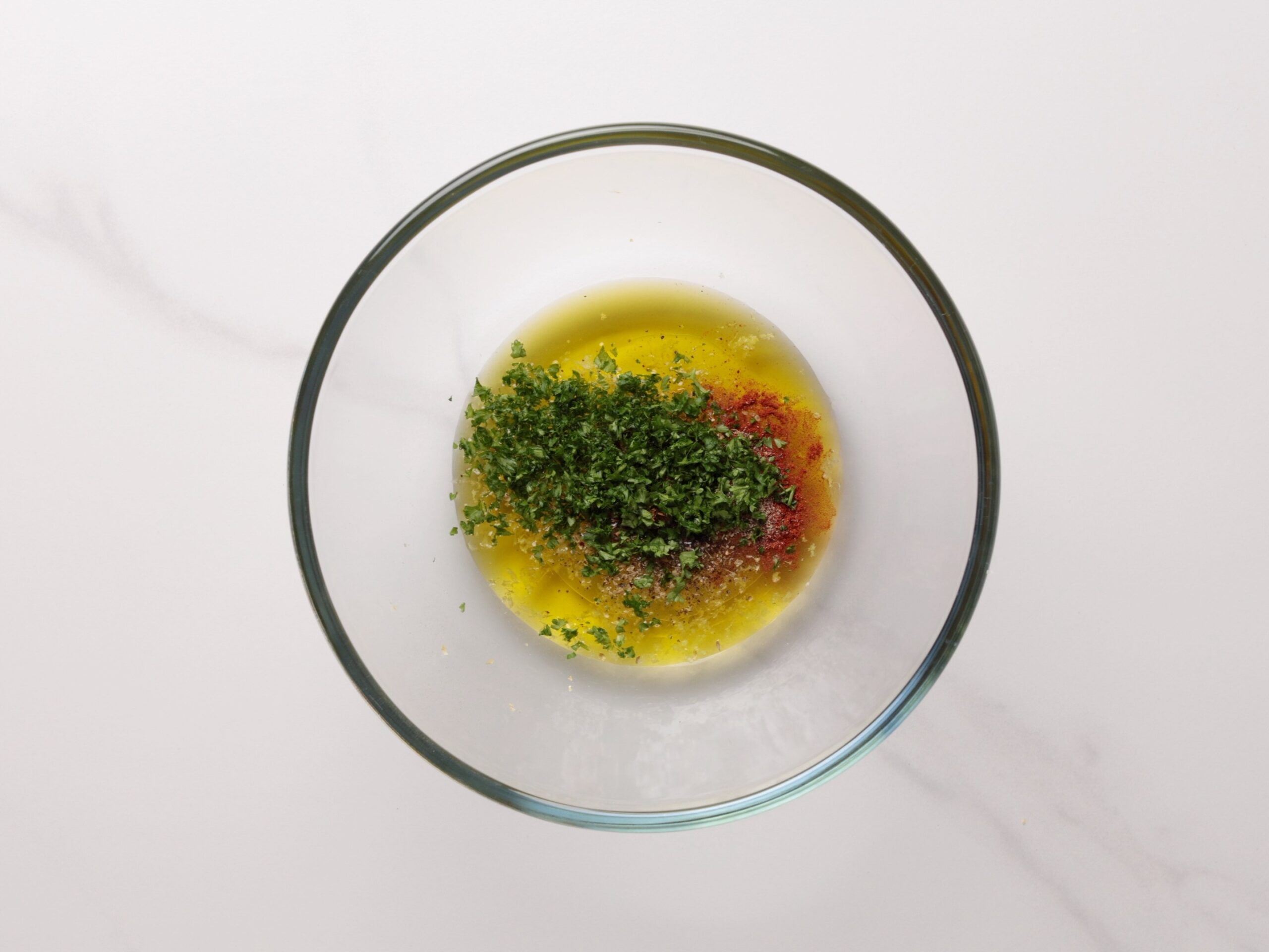 A glass bowl containing olive oil, finely chopped fresh herbs, and ground spices, sitting on a white marble surface.
