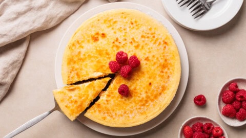 Crème Brûlée Cheesecake A cheesecake with a golden brulee top is garnished with fresh raspberries. One slice is being lifted with a spatula. Nearby are plates, forks, and bowls filled with more raspberries.