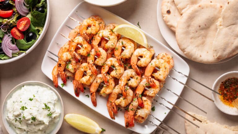 Grilled shrimp skewers on a white plate with lemon wedges, served alongside pita bread, a bowl of tzatziki, a salad with olives and cherry tomatoes, and a small dish of spices.