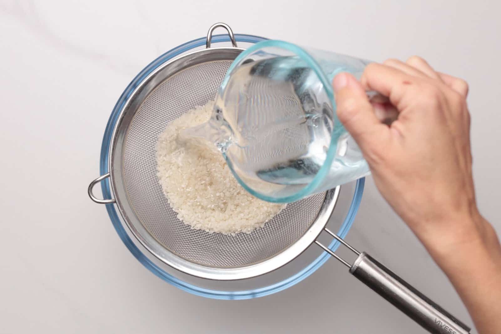 A hand pours water from a measuring cup over white rice in a metal strainer set above a blue bowl, rinsing the rice.