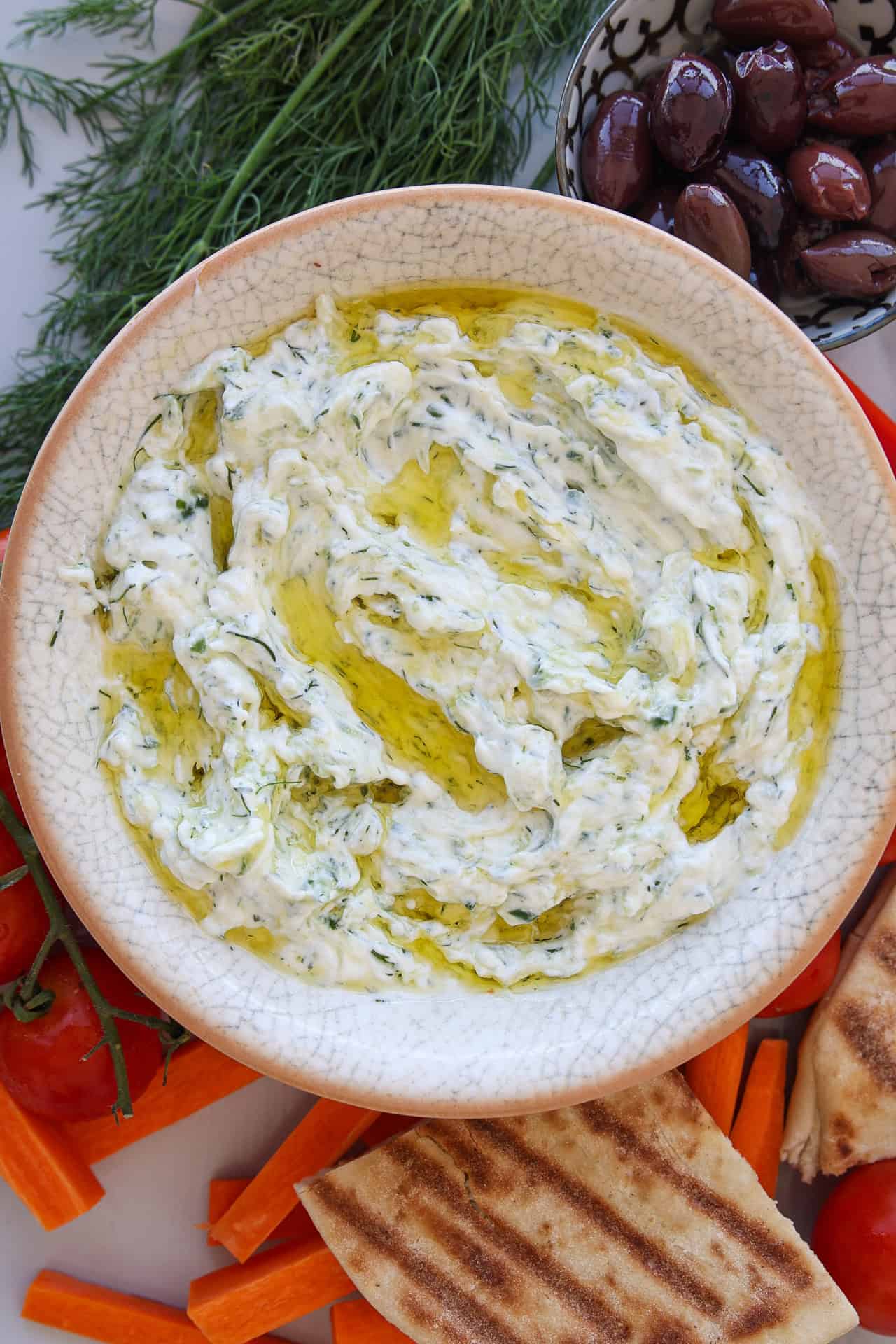 A bowl of creamy tzatziki dip drizzled with olive oil, surrounded by pita bread, carrot sticks, tomatoes, fresh dill, and a small bowl of black olives.