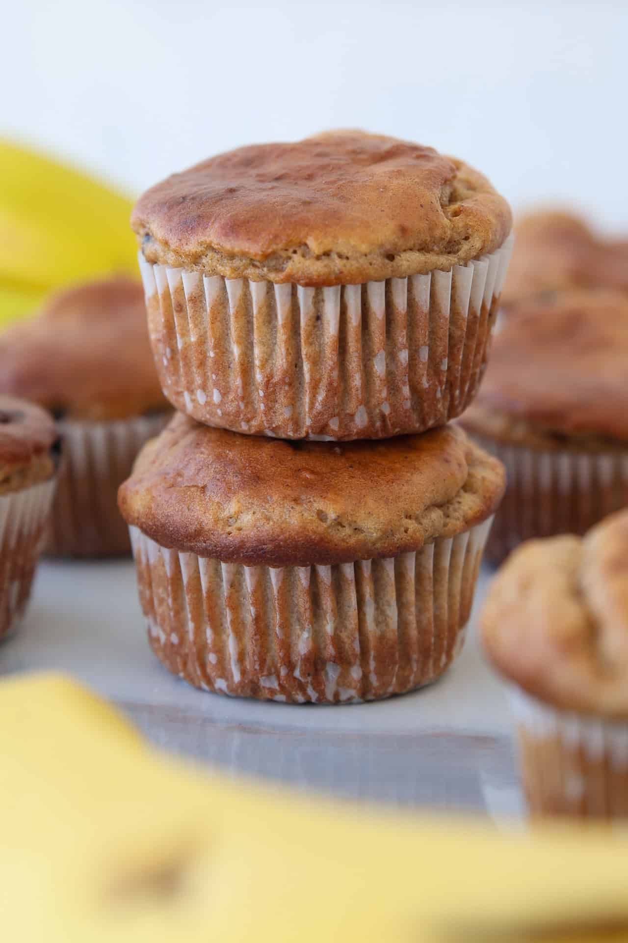 Two high-protein golden-brown banana cottage cheese muffins stacked on top of each other, with more muffins and blurred yellow bananas in the background.