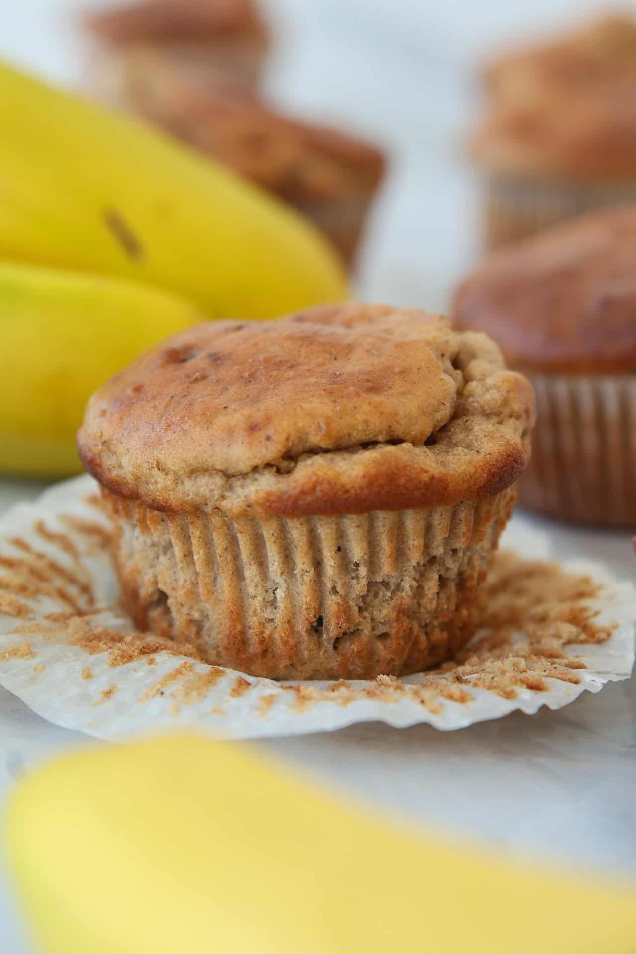 A banana protein muffin sits on a white paper liner with bananas and more muffins blurred in the background.