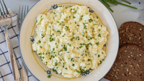 Healthy Cottage Cheese Egg Salad (High Protein & No Mayo) A plate of creamy cottage cheese egg salad garnished with chopped chives sits next to slices of brown seeded bread, two forks, and a blue-and-white checkered napkin.