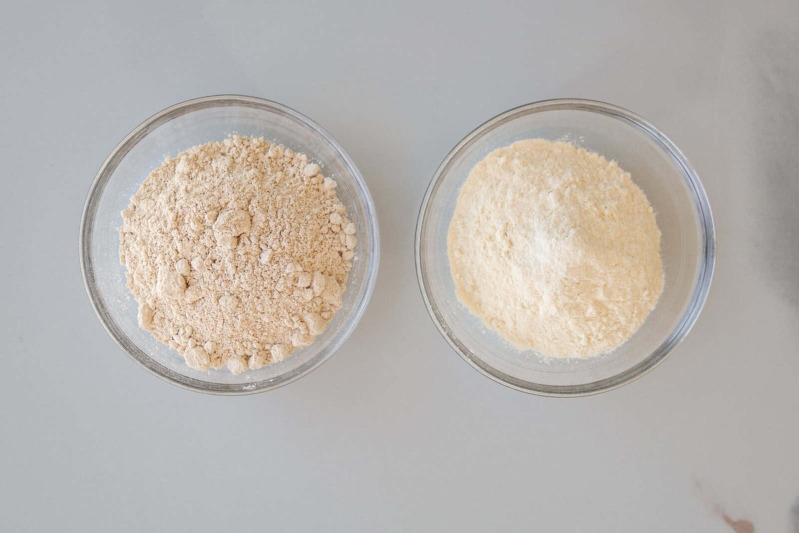 Two glass bowls sit side by side on a light surface. The bowl on the left contains Whole Wheat Flour, while the bowl on the right holds All Purpose Flour.