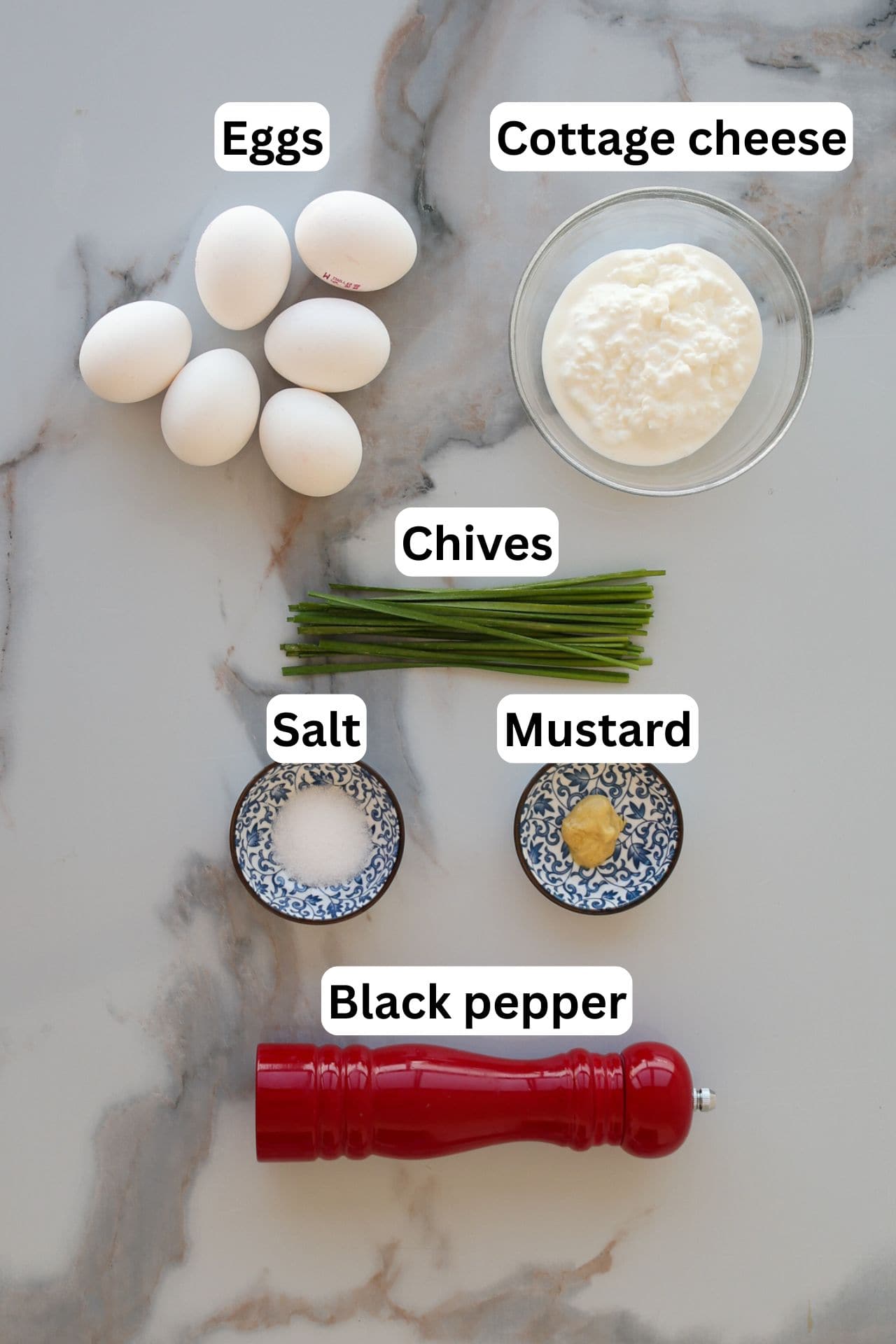 Six eggs, a bowl of cottage cheese, a bunch of chives, small dishes of salt and mustard, and a red pepper grinder, all arranged on a white marble surface with each item labeled.