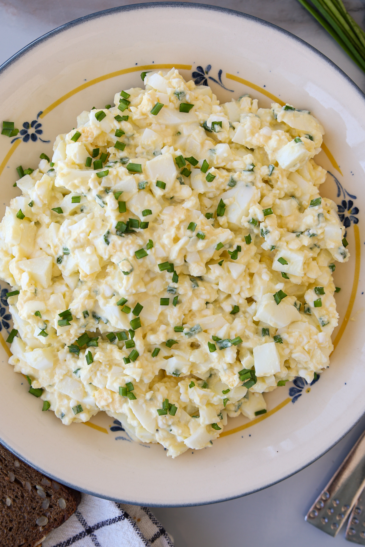 A bowl of creamy cottage cheese egg salad garnished with chopped chives, served in a decorative dish on a light surface with a patterned cloth and a slice of brown bread nearby.