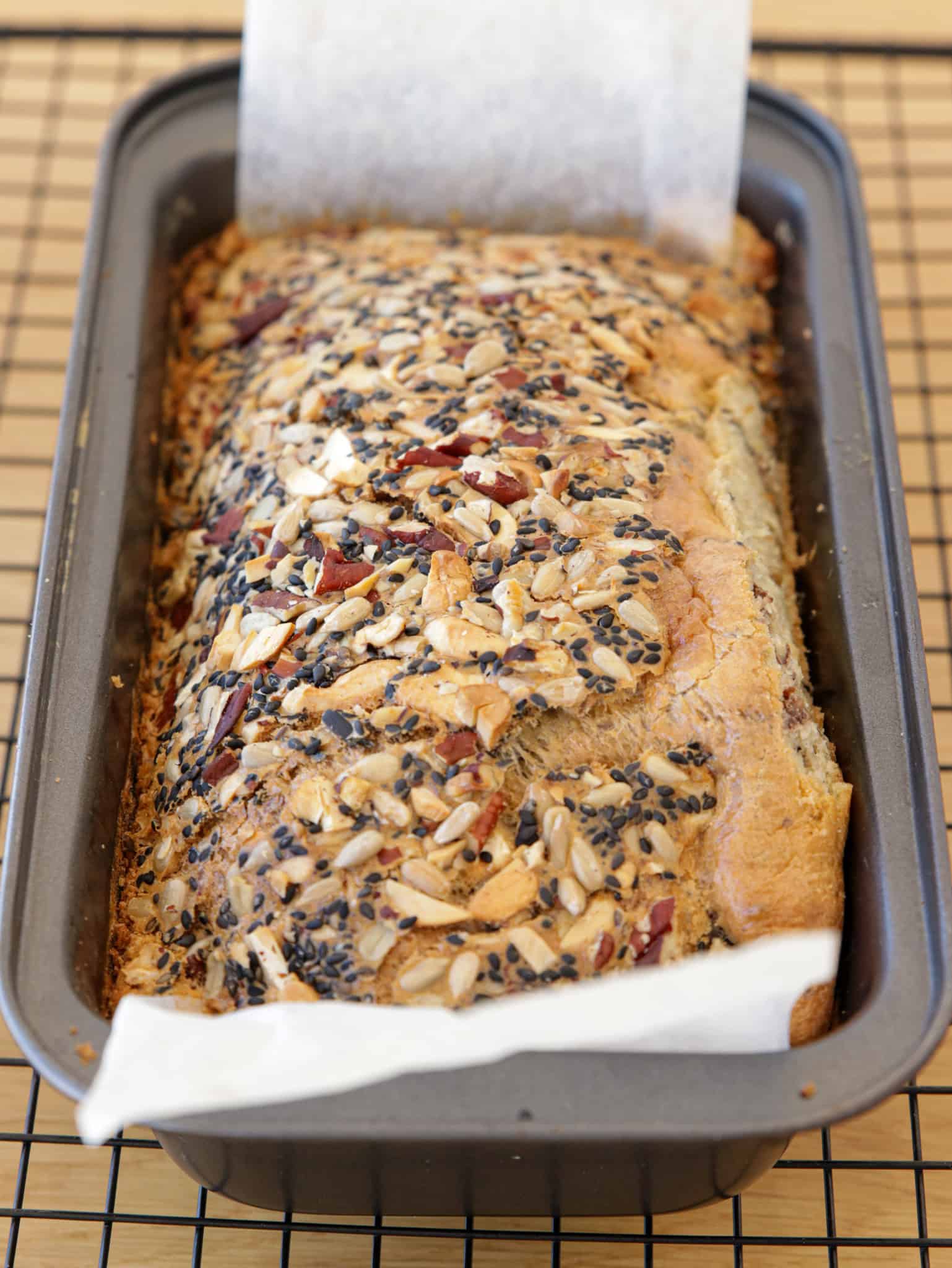 A flourless tahini bread topped with seeds and nuts sits in a parchment-lined metal baking pan on a cooling rack.
