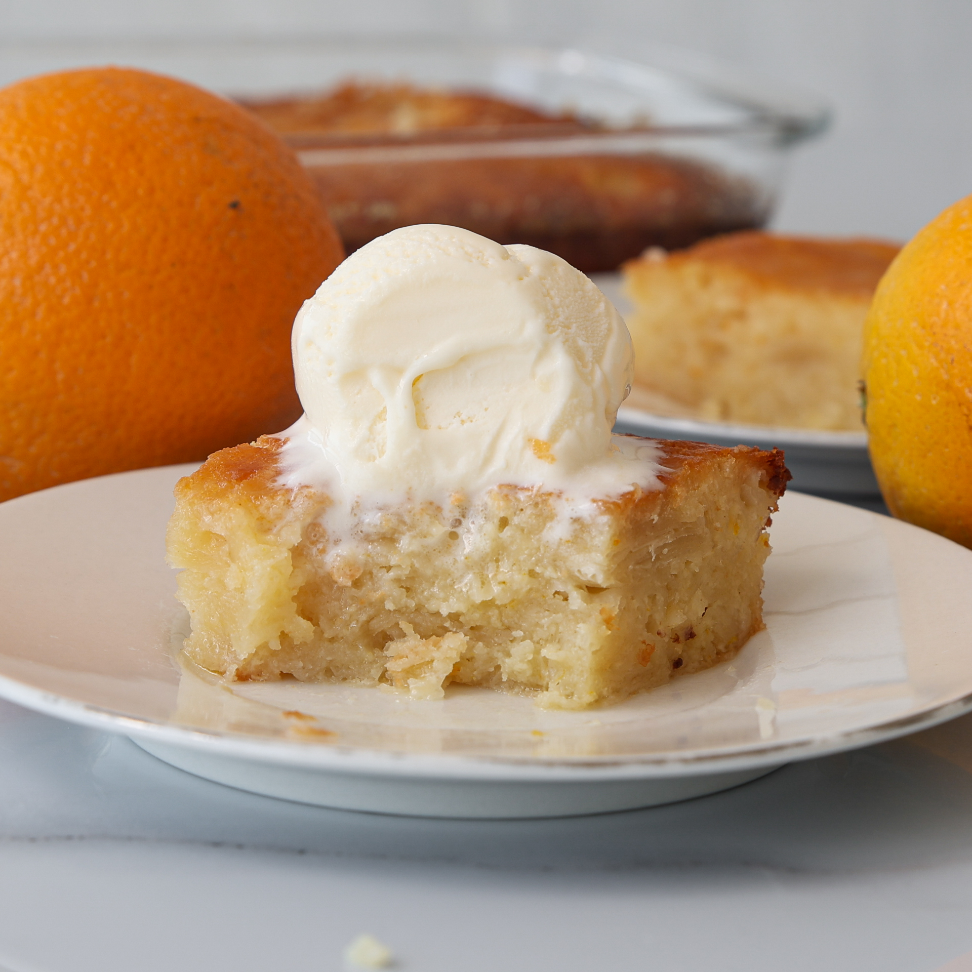 A square slice of moist portokalopita cake topped with a scoop of vanilla ice cream sits on a white plate. Whole oranges and more cake are visible in the background.
