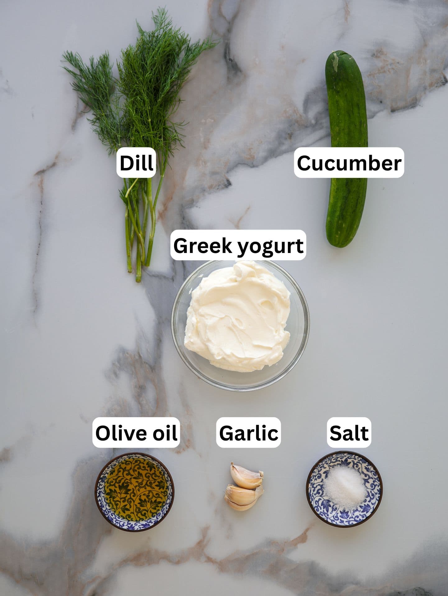 Overhead view of labeled ingredients for tzatziki: fresh dill, a cucumber, a bowl of Greek yogurt, a dish of olive oil, garlic cloves, and a dish of salt, all arranged on a marble surface.
