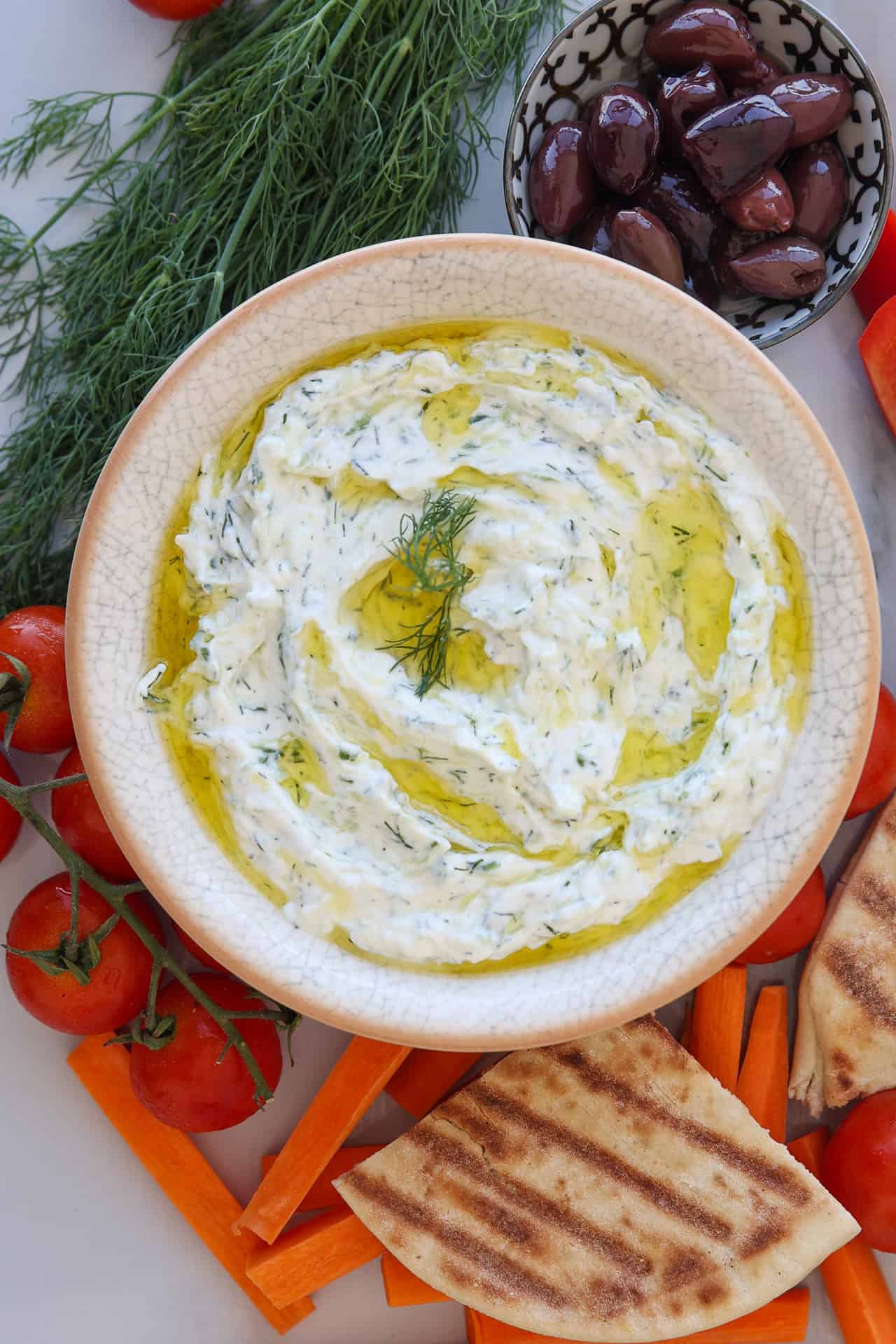 A bowl of creamy tzatziki dip garnished with dill and olive oil, surrounded by cherry tomatoes, olives, pita bread, and carrot sticks, on a white surface.