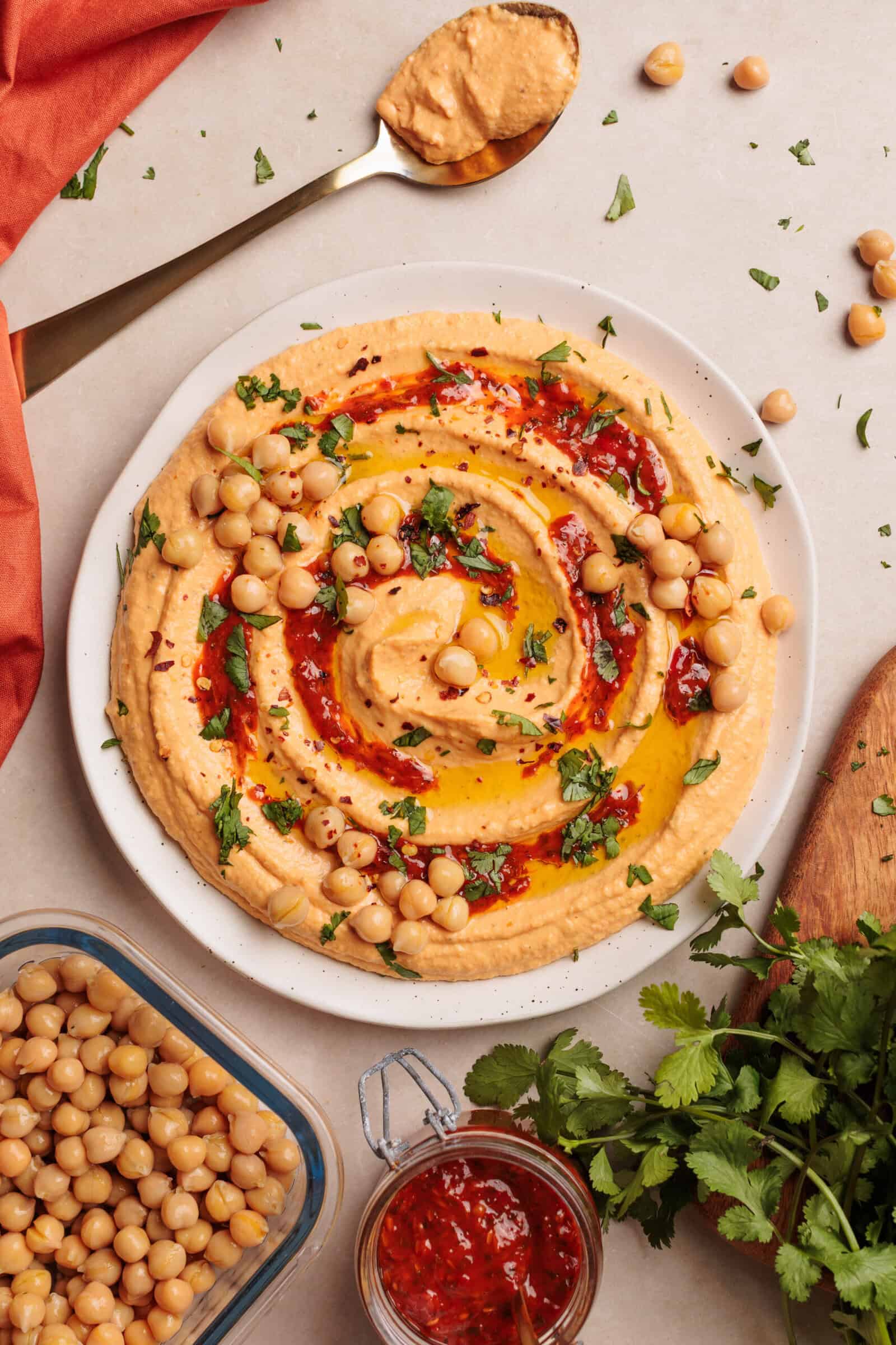 A plate of harissa hummus drizzled with olive oil, harissa paste, and garnished with chickpeas and chopped herbs. Chickpeas, a spoon with hummus, chili sauce, and fresh cilantro are arranged around the plate.
