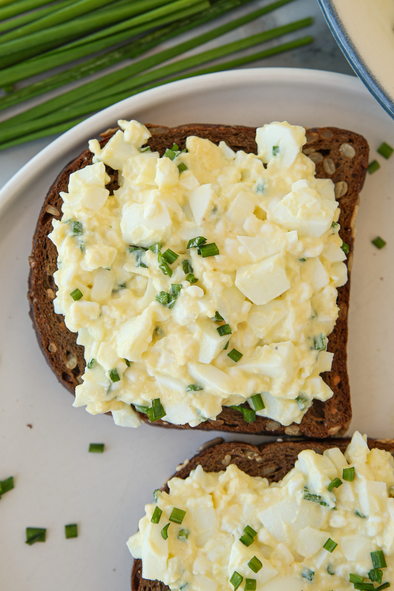 Two slices of dark bread topped with healthy cottage cheese egg salad and chopped chives on a white plate, with fresh chives in the background.