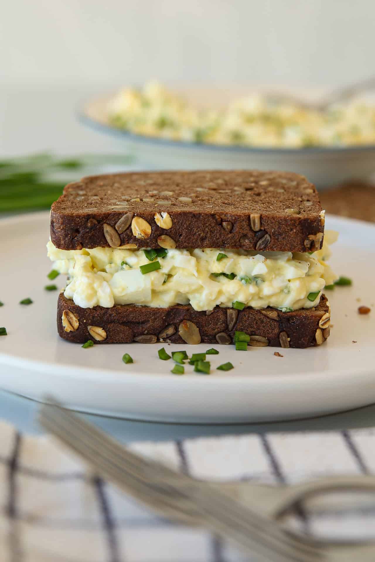 Healthy high-protein egg salad sandwich on multigrain bread, garnished with chopped chives, sits on a white plate with a fork and cloth nearby. A bowl of egg salad is blurred in the background.