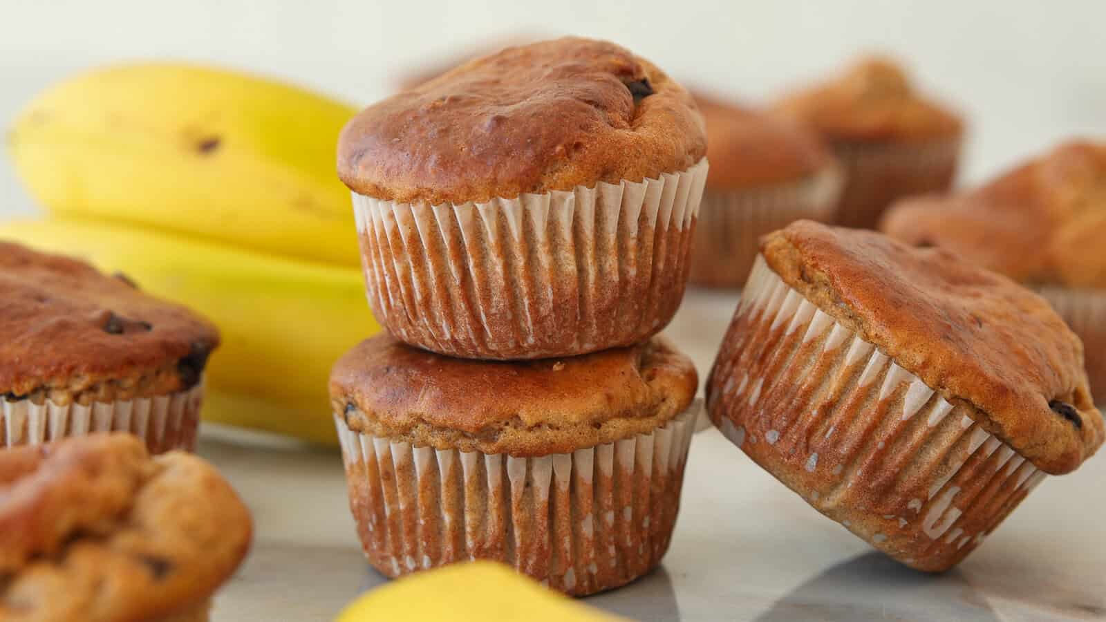 Two banana cottage cheese muffins are stacked in front of other muffins, with yellow bananas in the background. The muffins have golden-brown tops and are wrapped in white paper liners.