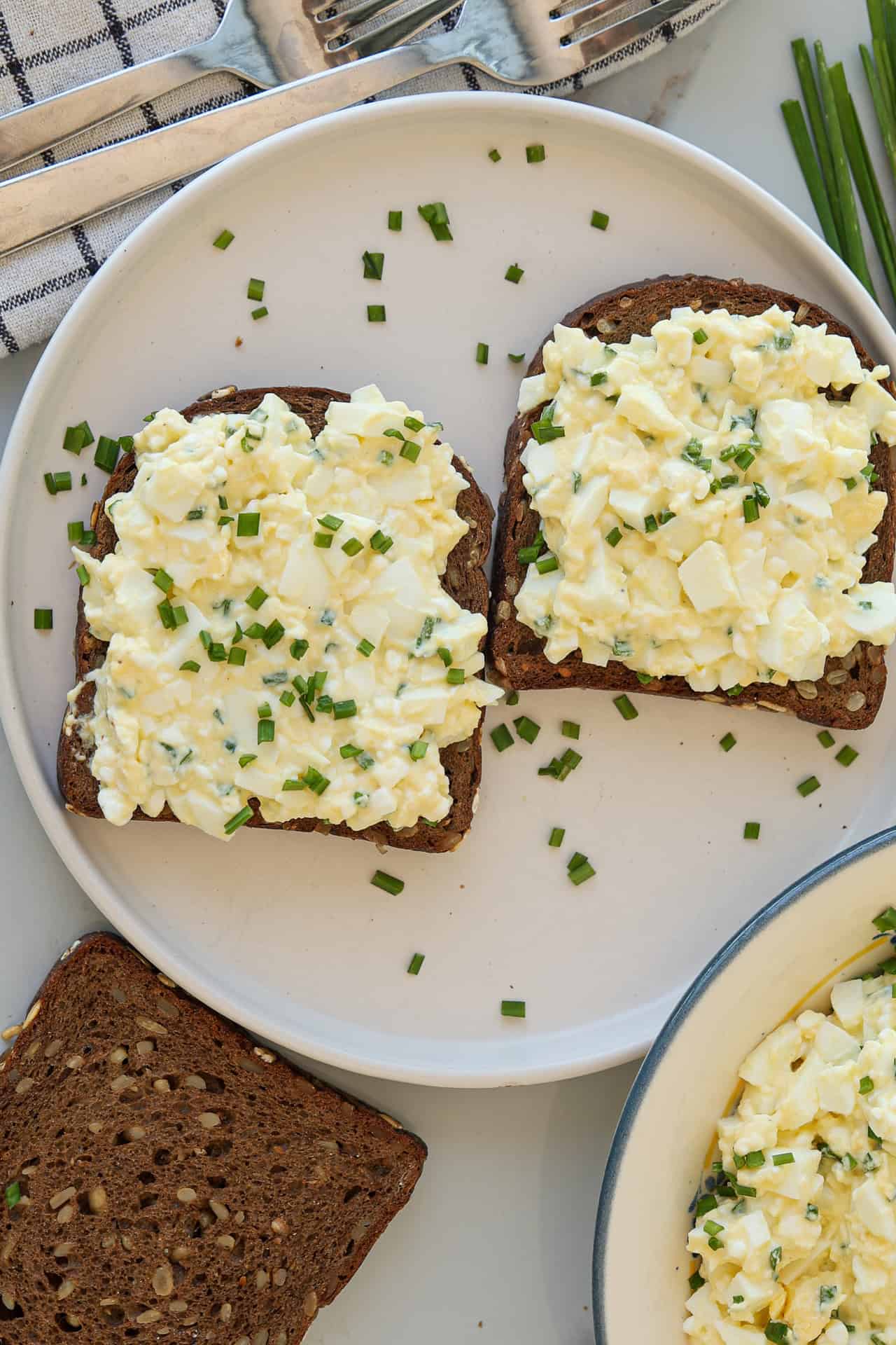 Two slices of dark bread topped with high-protein egg salad and chopped chives are on a white plate, with a third plain slice and a bowl of egg salad nearby.