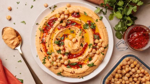Creamy Harissa Hummus Recipe A plate of harissa hummus topped with chickpeas, olive oil, chopped parsley, and homemade harissa sauce, surrounded by a spoon, fresh cilantro, extra chickpeas, and a small jar of harissa paste on a light surface.