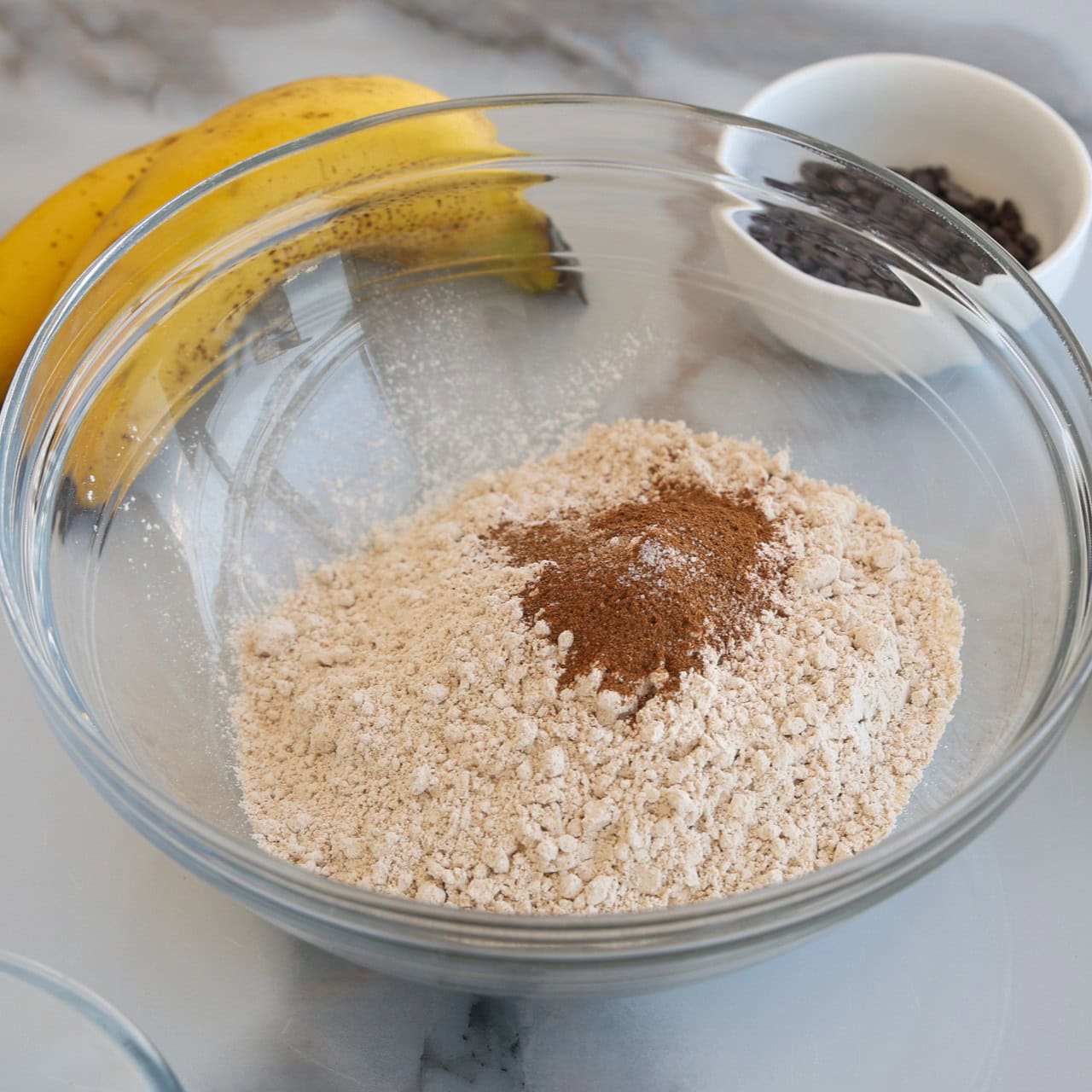 A clear bowl with flour and cinnamon sits on a counter near two bananas and a small white bowl filled with chocolate chips.