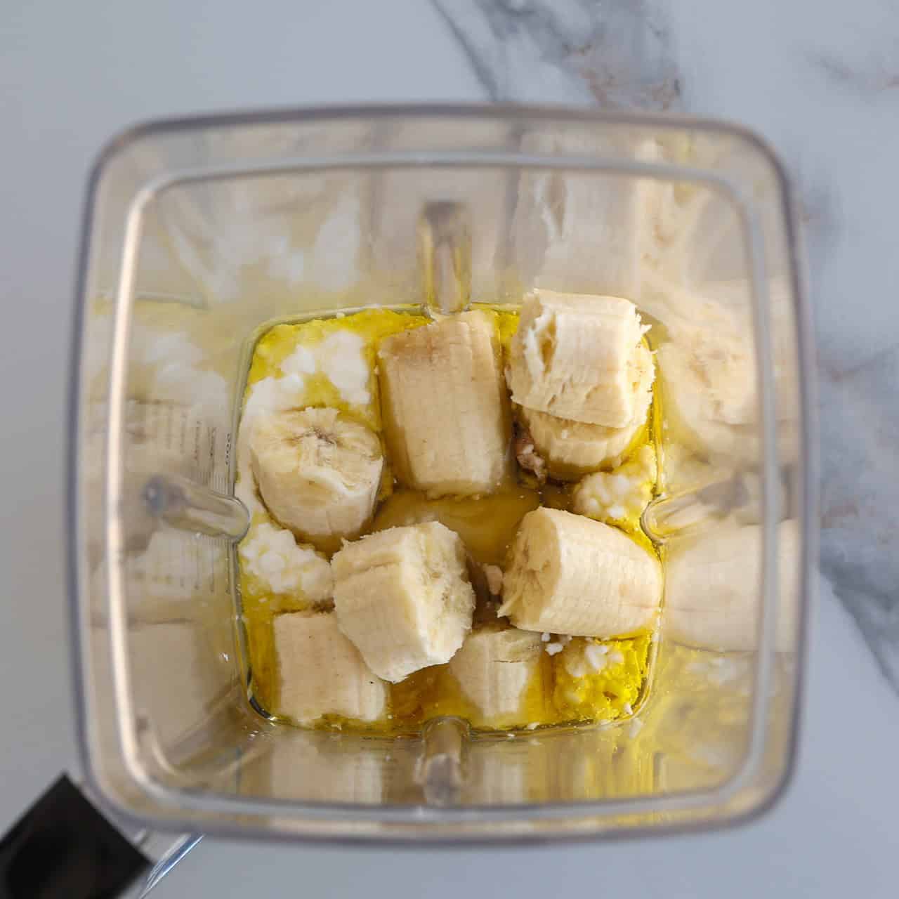 Overhead view of a blender containing chunks of banana, cottage cheese, olive oil, and vanilla on a white marble countertop.